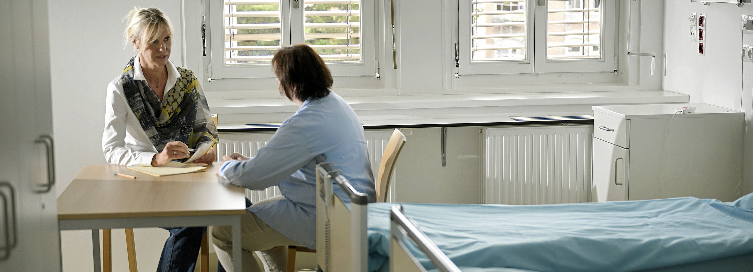 Vertretungsnetz Patientenanwältin im Gespräch mit einer Patientin an eine Tisch im Krankenhauszimmer, daneben ein Bett