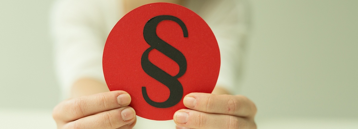 Foto: Zwei Hände halten ein rundes, rotes Schild in die Kamera, darauf ist ein Paragraphensymbol abgebildet