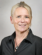 Porträtfoto einer lächelnden Frau mit blondem, zurückgebundenem Haar, schwarzer Bluse und schlichtem Anhänger vor neutralgrauem Hintergrund.