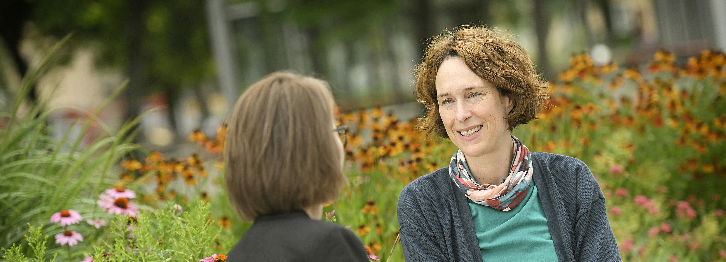 Ehrenamtliche Mitarbeiter:innen Foto: Erwachsenenvertreterin und Klientin im Gespräch auf einer Bank im öffentlichen Raum, dahinter Blumen