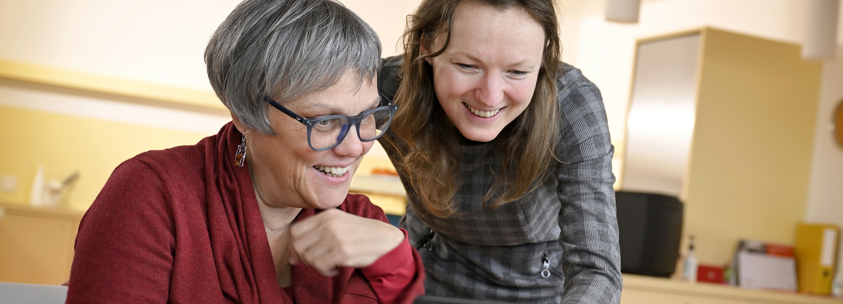 Vertretungsnetz Foto: Zwei Frauen schauen lächelnd auf einen Laptop-Bildschirm. Eine Frau sitzt und tippt auf dem Laptop, während die andere Frau dahinter steht und sich über den Bildschirm beugt.